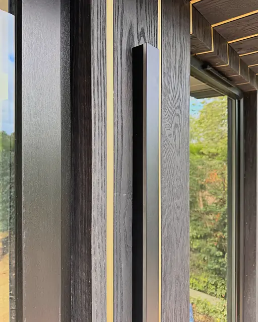 Close up of charred-black timber cladding beside a glazed door, a slim gold reveal running up the board and along the timber soffit.