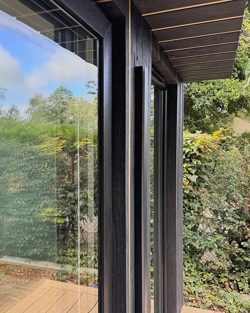 Looking out past the corner of a black timber garden studio: charred-black boards with a slim gold reveal, a full-height glass slider and the garden beyond the deck.