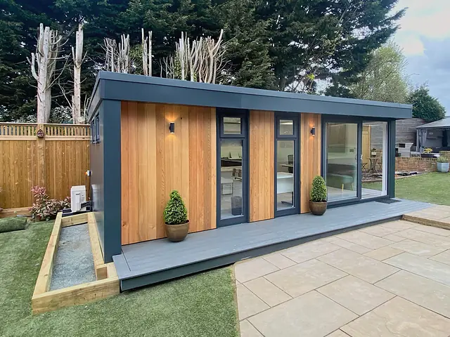 A garden office in vertical cedar boards framed in anthracite, on a grey composite deck with two clipped topiary pots, beside a sandstone patio and a timber raised bed.