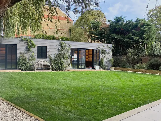 A long garden studio in pale grey boards with black-framed glazed doors and climbing plants, seen across a wide lawn under a willow.