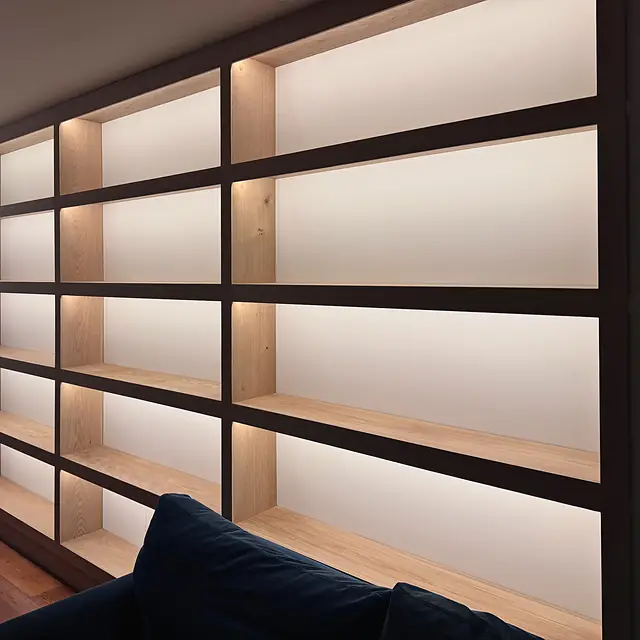 A wall of oak shelving framed in dark timber, every bay lit from behind, glowing in the evening.