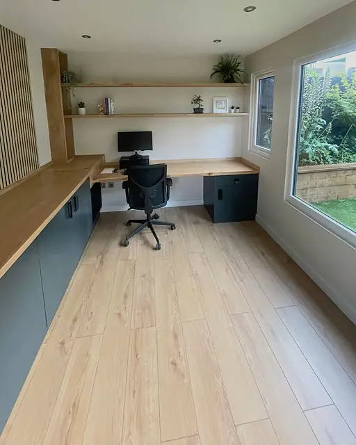 Inside a garden office: a long oak desk over dark cabinets, oak floating shelves above it, an oak slatted return and a large window onto the garden.