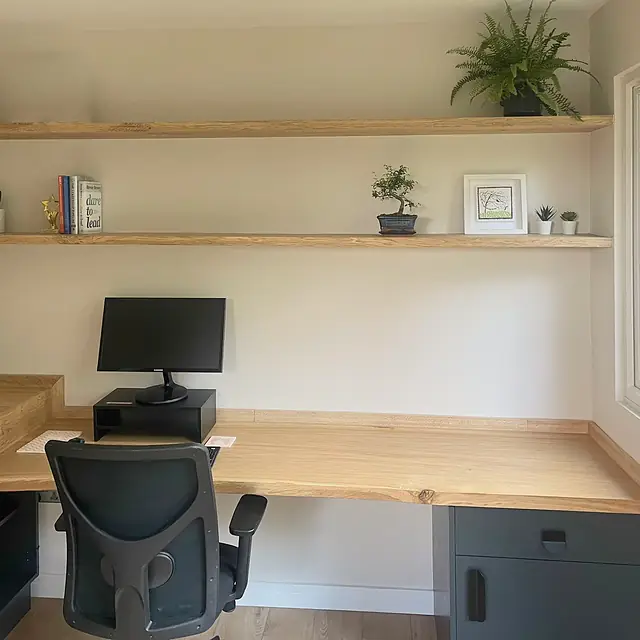 A garden office corner: a solid oak desk over a dark grey cabinet, two live-edge oak shelves above it, a fern and a bonsai on the shelves.