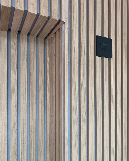 Oak slat panelling on a dark backing, close, where the slats turn the edge of an opening, with a black switch plate set into the wall.