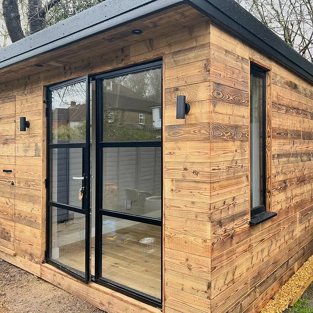A small garden studio clad in weathered reclaimed timber boards, with slim black-framed glazed doors, a side window and black wall lights.