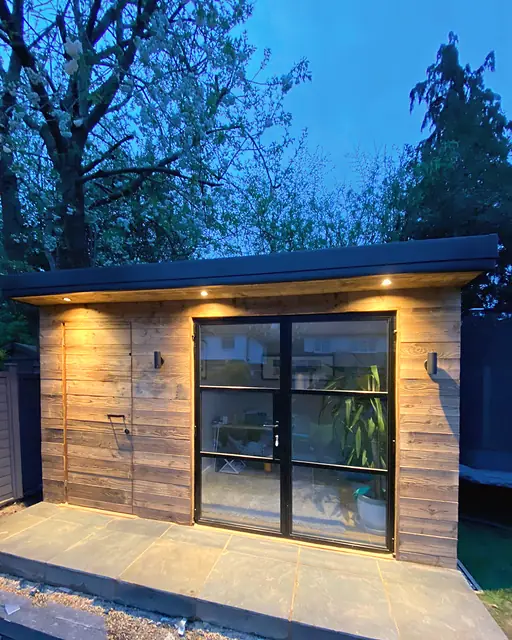 A garden studio clad in weathered reclaimed boards at dusk, the soffit downlights and wall lights on above slim black-framed glazed doors.
