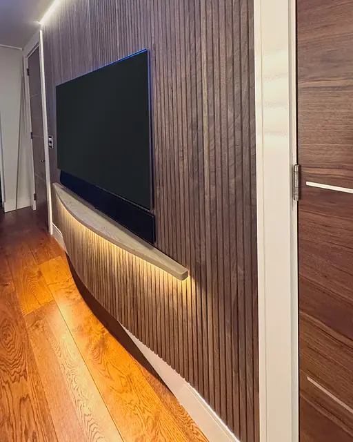 A dark walnut slat wall holding a television, lit by a hidden strip along the ceiling and a glowing curved plinth over an oak floor.