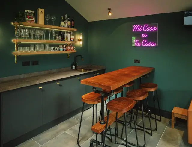 A bar corner with open shelving, a green-painted counter, an oak high table and stools.