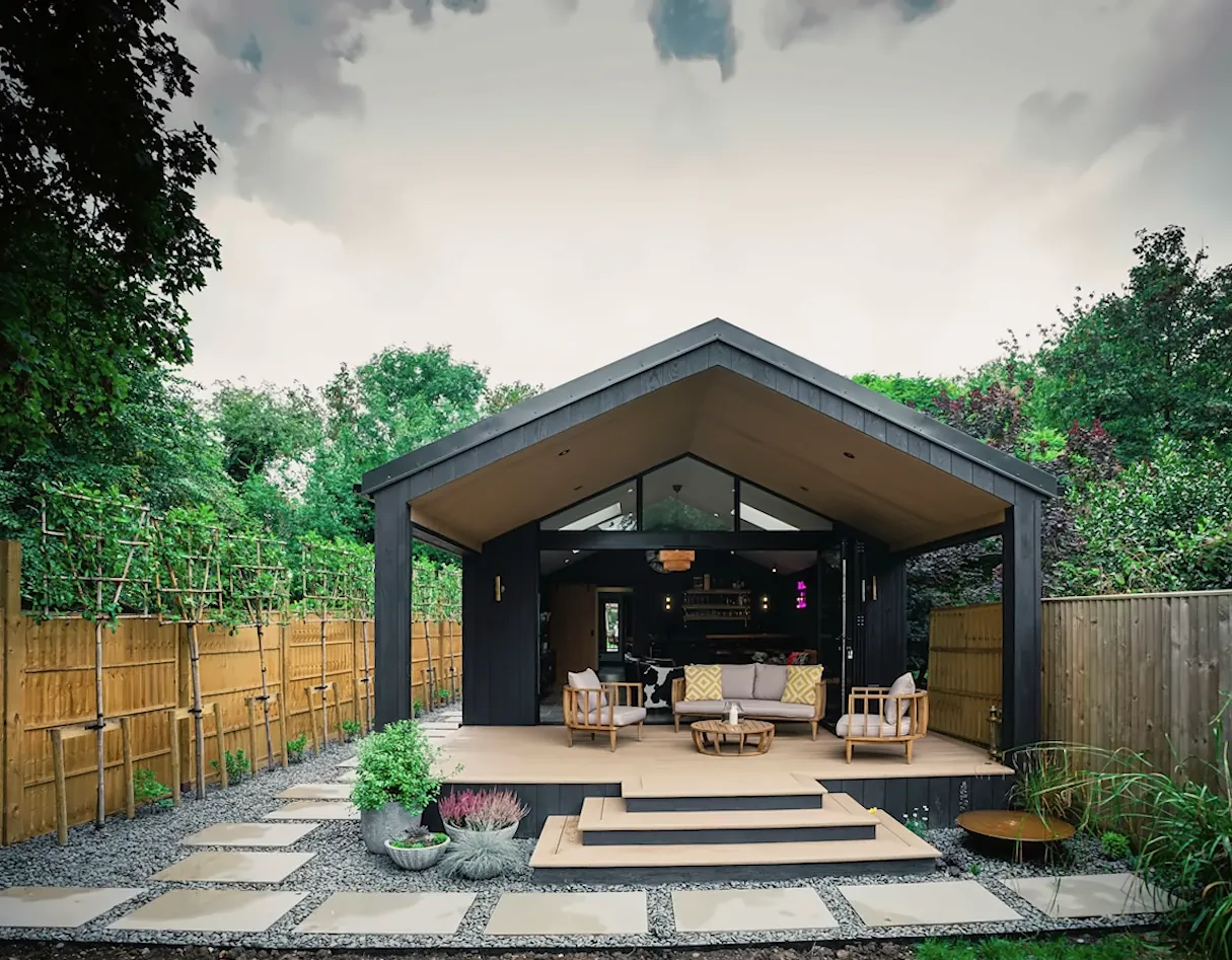 A black timber pavilion built by KF Crafts, with an open gable end, an outdoor lounge inside and wide steps down to a stepping-stone path.