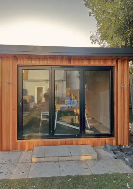 A cedar-clad garden room in low sun with black-framed glazed doors fitted, a slab step in front and tools still inside.