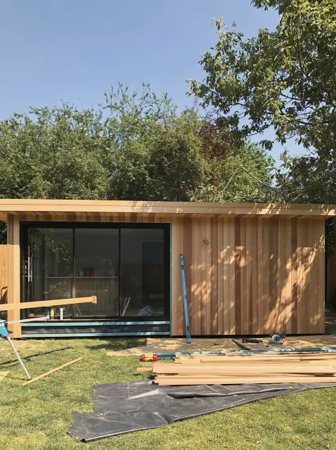 A long garden room part-way through cladding: cedar boards going on, black-framed glazing already in and more boards stacked on the lawn.