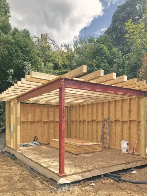A red steel portal frame standing inside a timber-framed building part-way through its build.