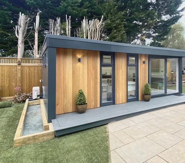 A garden office in vertical cedar boards framed in anthracite, with glass doors onto a patio.