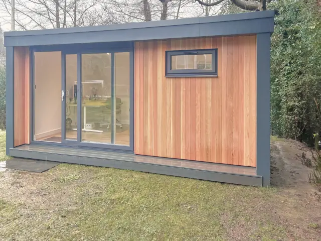 A garden office in vertical cedar boards framed in anthracite, with sliding glass doors and a small high window, on a lawn among bare winter trees.