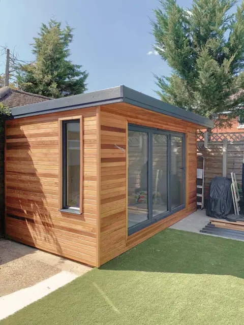 A cube of a garden room in horizontal cedar boards with a black fascia and black-framed bifold doors, on an artificial lawn.