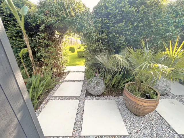 Large pale stone slabs set in gravel, leading between dense planting past a palm in a clay pot and wire spheres.
