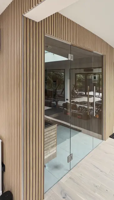 A frameless glass door set into a wall of pale timber slats, opening onto a bright room under a rooflight.