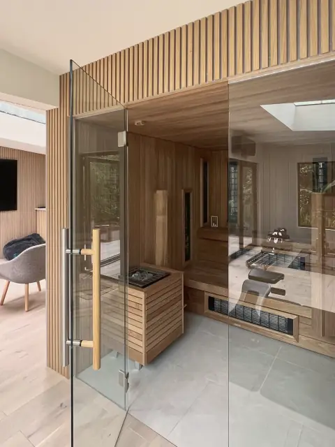 A sauna with a full glass front and door, lined in pale timber, set into a room with slatted walls and an oak floor.