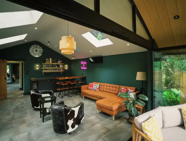 A vaulted lounge under rooflights: a leather corner sofa, a bar along a dark green wall and glass doors to the garden.