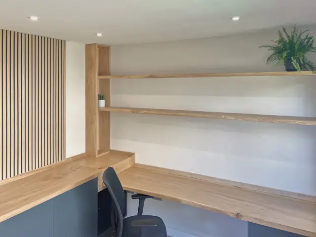 A corner home office: a live-edge oak worktop over dark cabinets, two floating oak shelves on an oak upright and a slatted timber wall.
