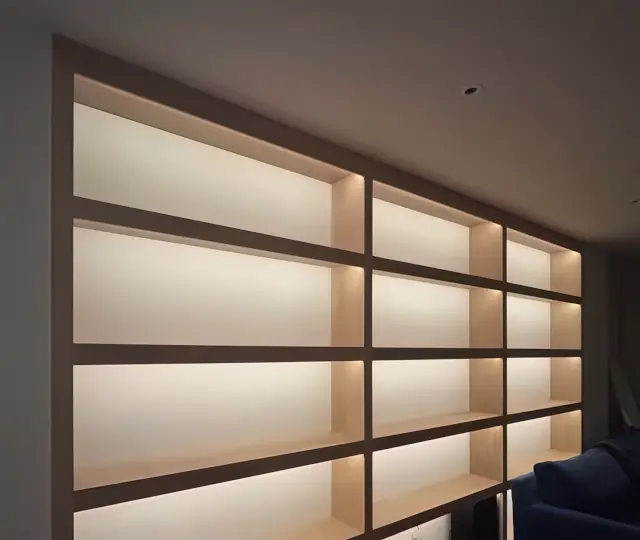 A wall of oak shelving, each bay lit from within, photographed at night.