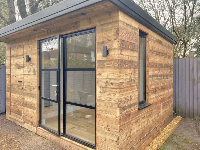 A small garden studio clad in weathered reclaimed boards, with black-framed glass doors and a wall light.