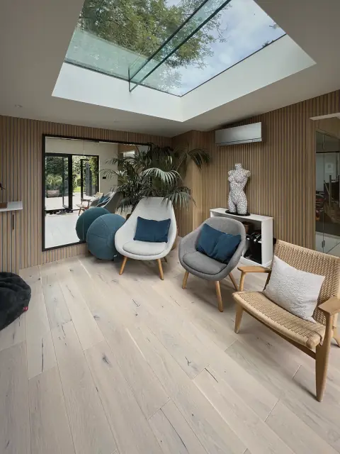 A bright lounge under a large flat rooflight, with an oak floor, slatted walls and armchairs.