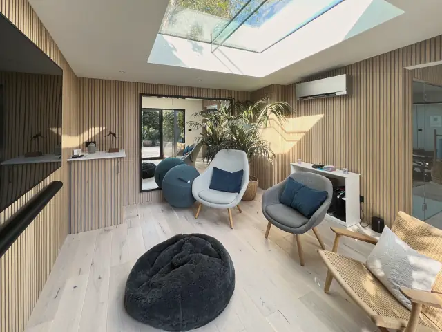 A lounge lined in pale oak slats under a walk-on rooflight, in warm low sun: armchairs, a rope-seat chair, a bean bag, a palm and a large wall mirror.
