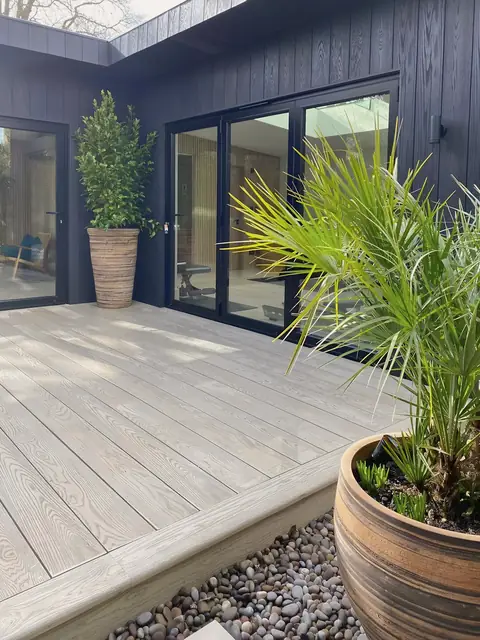 A studio in black vertical boards wrapping around a pale composite deck, with folding glass doors, a potted bay tree and a palm in a clay pot.