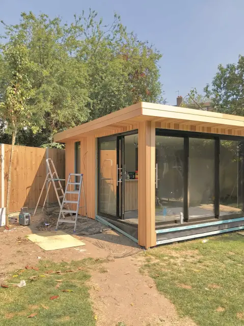 A cedar-clad garden room with a deep cedar canopy and full-height glass, nearly finished, with stepladders still on site and the lawn still to be made good.