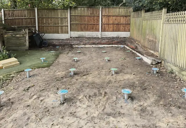 Rows of steel ground screws with blue caps set into a cleared patch of garden, ready for the floor frame.