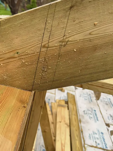 Close up of a treated timber rafter with pencil setting-out lines, seated on the frame below, with insulation boards stacked beyond.