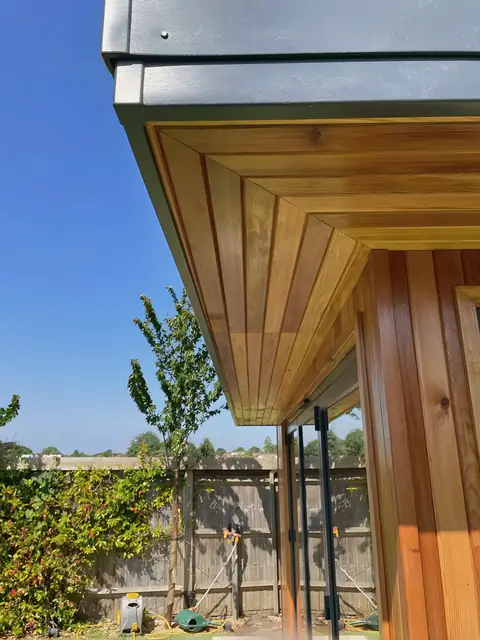Looking up at the corner of a garden room: cedar soffit boards meeting in a mitre under an anthracite fascia, against a blue sky.