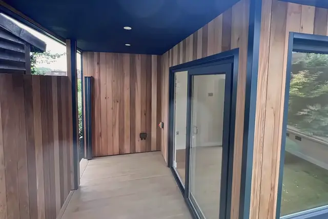 A garden room in vertical cedar boards with a deep covered porch, a sliding glass door and a large corner window.