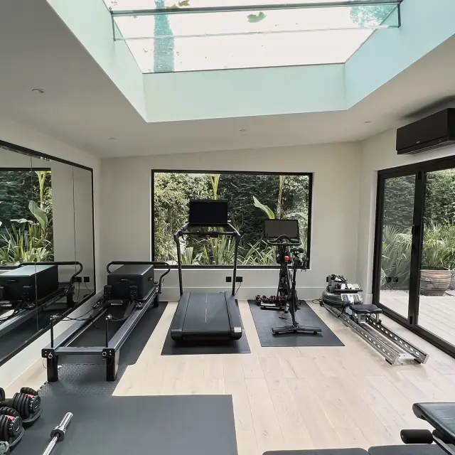 A garden gym under a large rooflight: a mirrored wall, a reformer, a treadmill, an exercise bike and a rower on a pale oak floor, with glass doors to a planted garden.