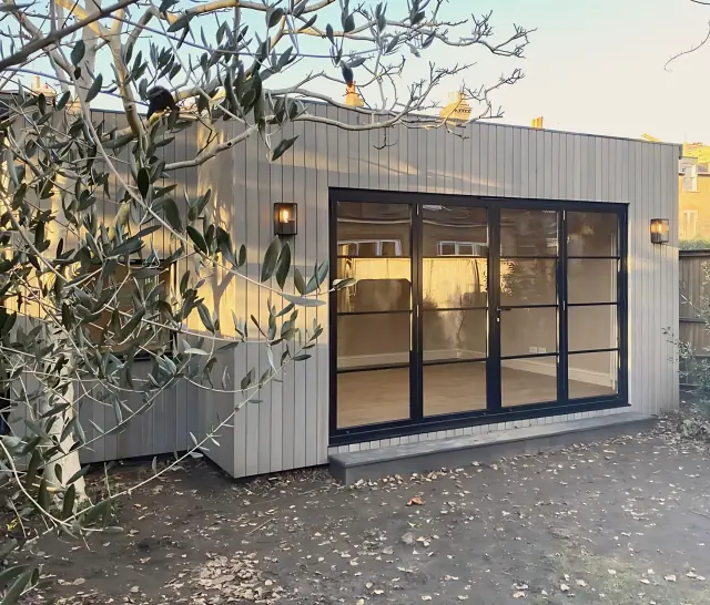 A garden studio in pale grey vertical boards with black-framed glazed doors and a wall lantern either side, in low evening sun behind an olive tree.
