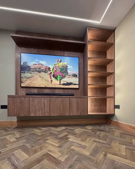 A timber media wall: a screen framed in dark veneer above a low cabinet, a tall run of open shelves lit from within, and a herringbone floor.