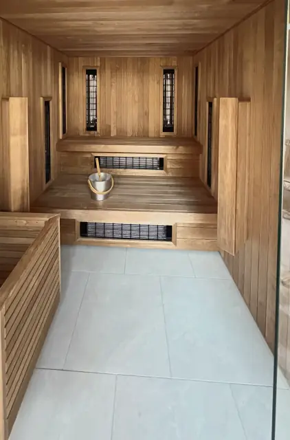Inside a timber-lined sauna, seen past its glass door: slatted benches, infrared heater panels in the walls, a bucket and ladle, and a pale tiled floor.