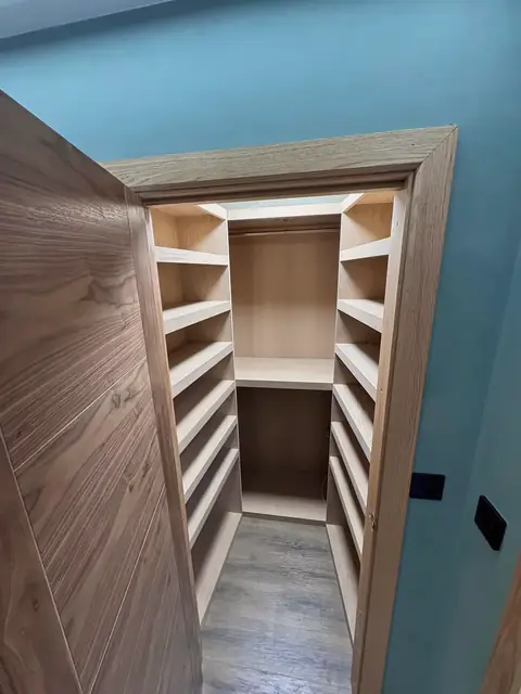 A dark veneered door opening onto a walk-in wardrobe lined with pale timber shelves, set in a teal-painted wall.