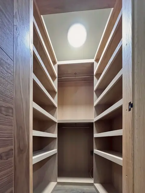 A walk-in wardrobe: pale timber shelving on both sides and at the back, with hanging space and a ceiling light, seen through a dark veneered doorway.
