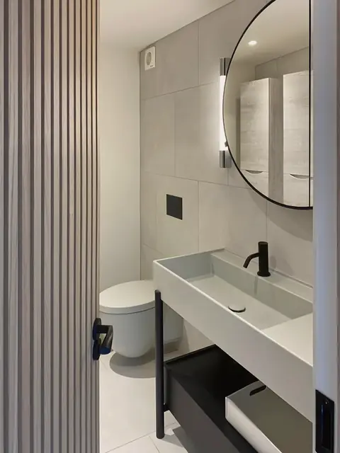 A washroom behind a door in a slatted wall: large pale tiles, a wall-hung WC, a trough basin on a black steel stand and a round black-framed mirror.