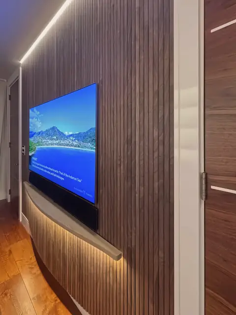 A dark walnut slat wall holding a television, lit by a strip of light along the ceiling and the floor.