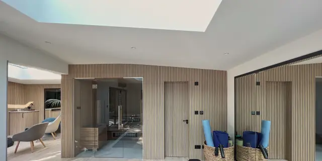 A wellness room under a large rooflight: walls of pale timber slats, a glass-fronted sauna, a flush door set into the slats and a mirrored wall reflecting the room.