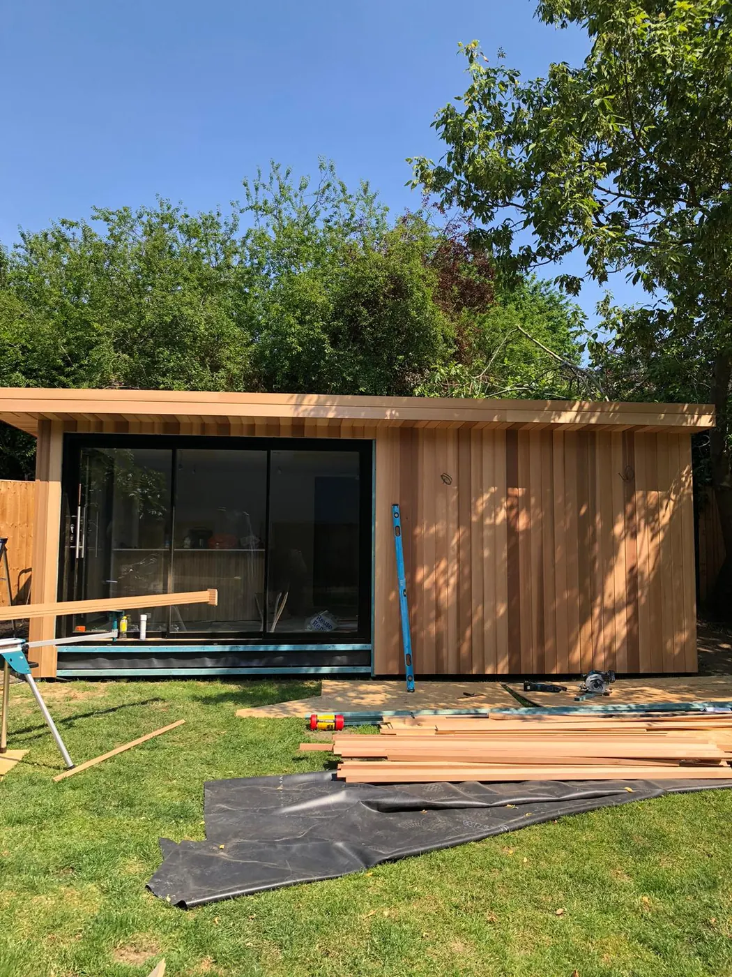 A garden room part-way through its build: frame up, cedar cladding going on, timber stacked on the lawn.